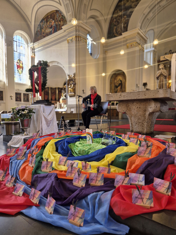 Bunte Stoffbahnen in Spirale auf dem Boden einer Kirche, darauf kleine Karten mit Windmühlenmotiven, im Hintergrund eine Person mit Akkordeon vor Altar und Kirchenfenstern.