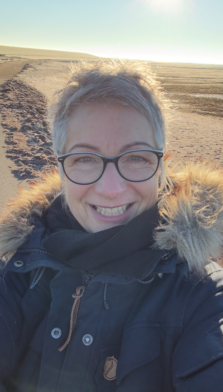 Angela Axmann mit dunklem Wintermantel mit Fellkapuze vor einer weiten, sonnigen Sandlandschaft.