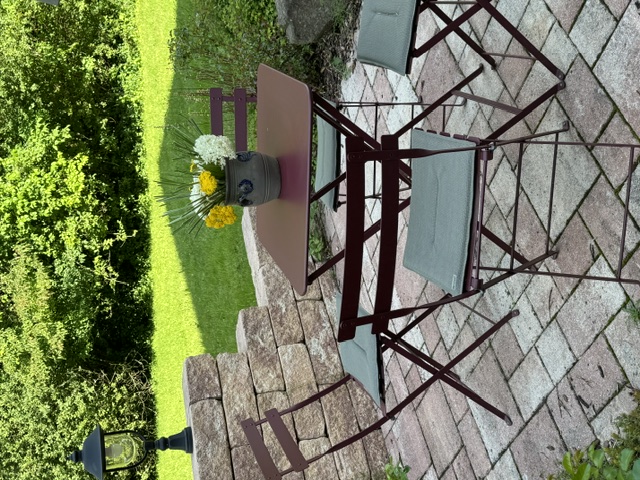 Garten mit gepflastertem Boden, einem runden Tisch mit Blumenvase und vier Stühlen mit Sitzkissen, umgeben von einer niedrigen Steinmauer und grünem Buschwerk