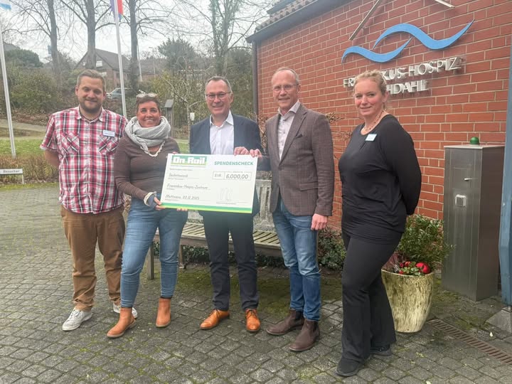Fünf Personen stehen vor einem Gebäude und halten gemeinsam einen großen Spendenscheck