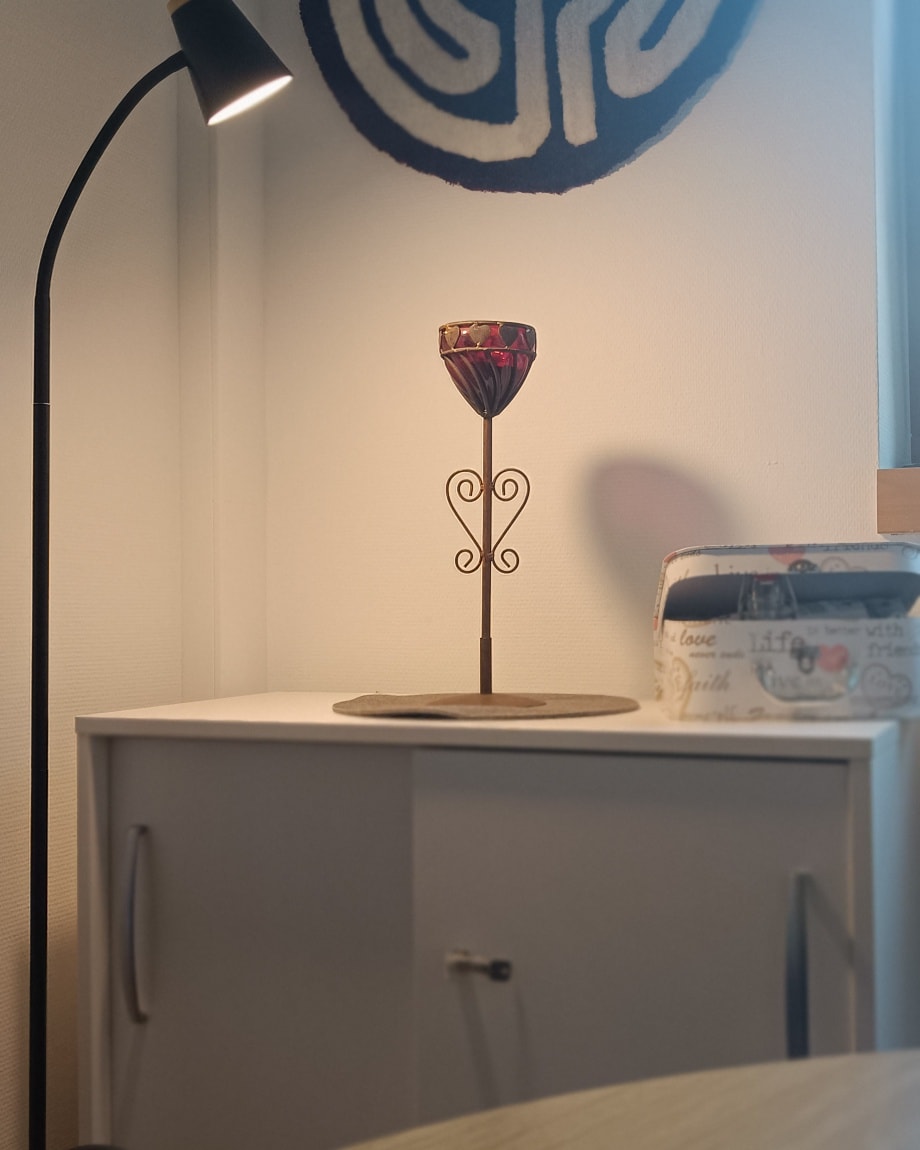 Kerzenhalter mit verziertem Stiel auf weißem Schrank, daneben weiße Box mit Schrift, links Stehlampe, Wand mit blau-weißem Wandbehang