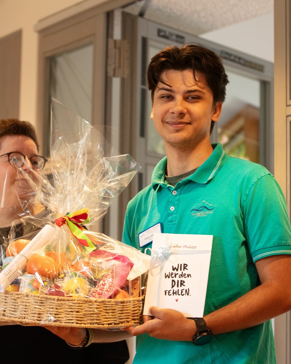 Mann in grünem Poloshirt hält einen Geschenkkorb mit bunten Lebensmitteln und eine Karte mit der Aufschrift 'WIR WERDEN DIR FEHLEN'.