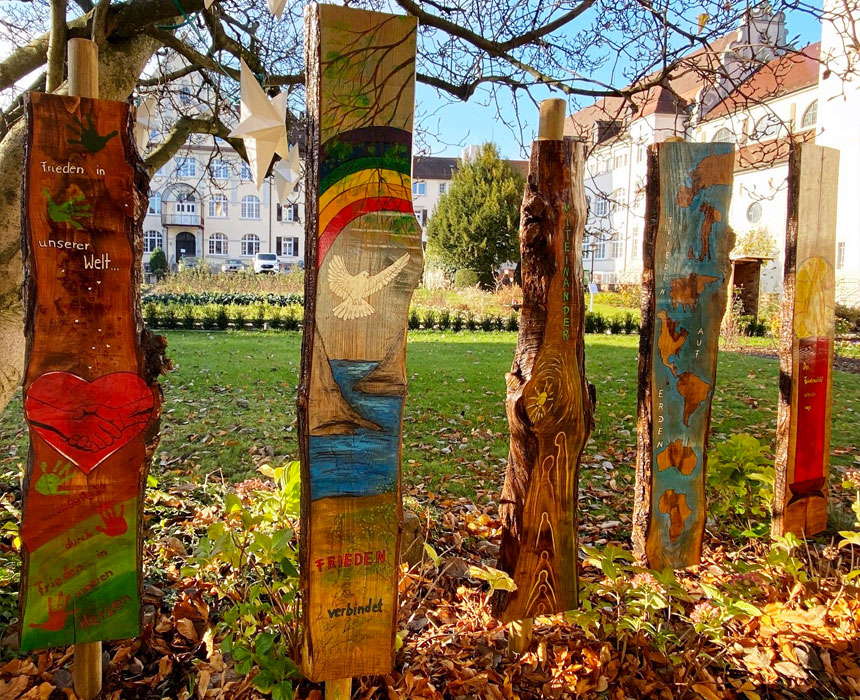 Fünf bemalte Holzpfähle mit Friedensthemen, darunter Herz, Taube, Regenbogen und Schrift, stehen im Herbstlaub auf einer Wiese vor Gebäuden.