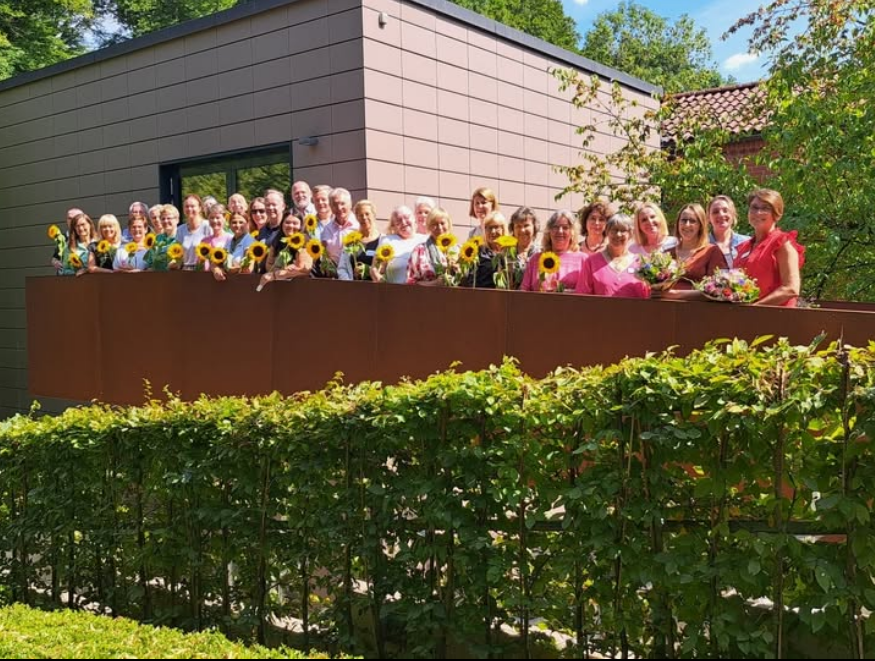 Gruppe von Menschen steht auf einer Terrasse vor modernem Gebäude, jede Person hält eine Sonnenblume oder einen Blumenstrauß.