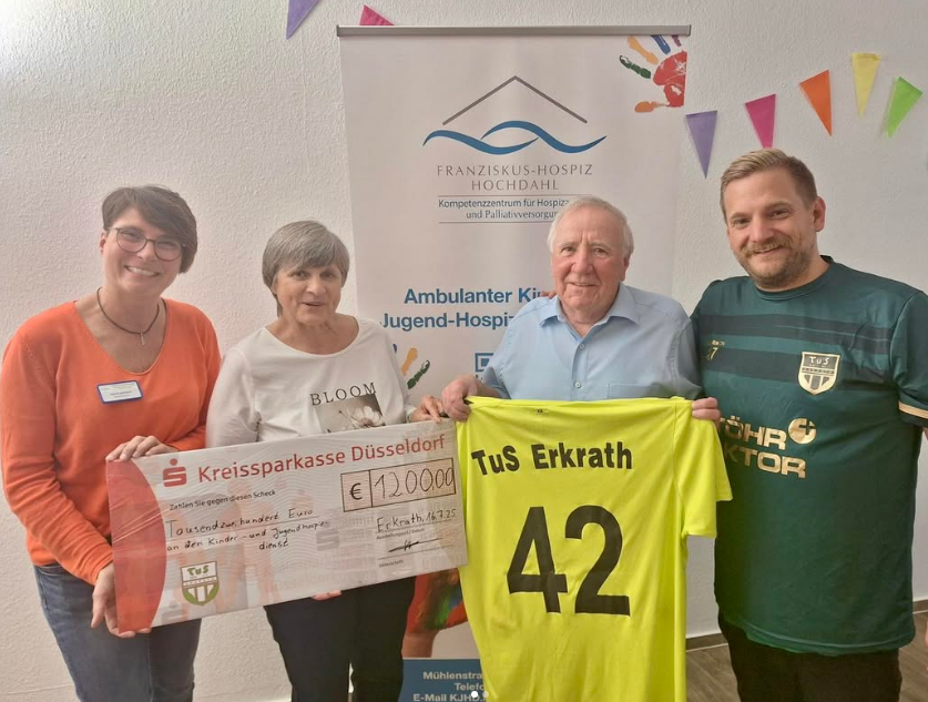 Vier Personen stehen vor einem Banner des Franziskus-Hospiz Hochdahl. Zwei halten einen großen Scheck über 12.000 Euro, zwei weitere halten ein gelbes Fußballtrikot mit der Aufschrift 'TuS Erkrath 42'.