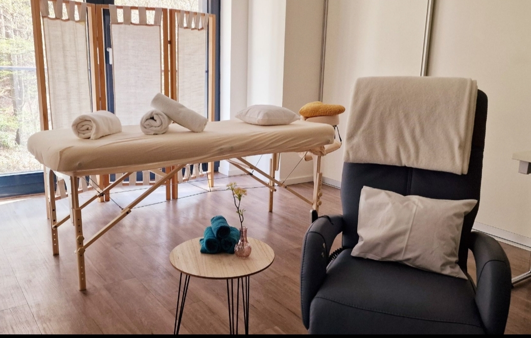 Massageliege mit weißen Handtüchern und Kissen in hellem Raum, daneben schwarzer Sessel mit Kissen und Decke, kleiner runder Tisch mit blauer Rolle und Vase.
