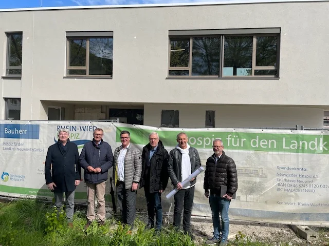 Sechs Männer in Jacken stehen vor einem Bauzaun mit Banner vor einem modernen Gebäude mit großen Fenstern.