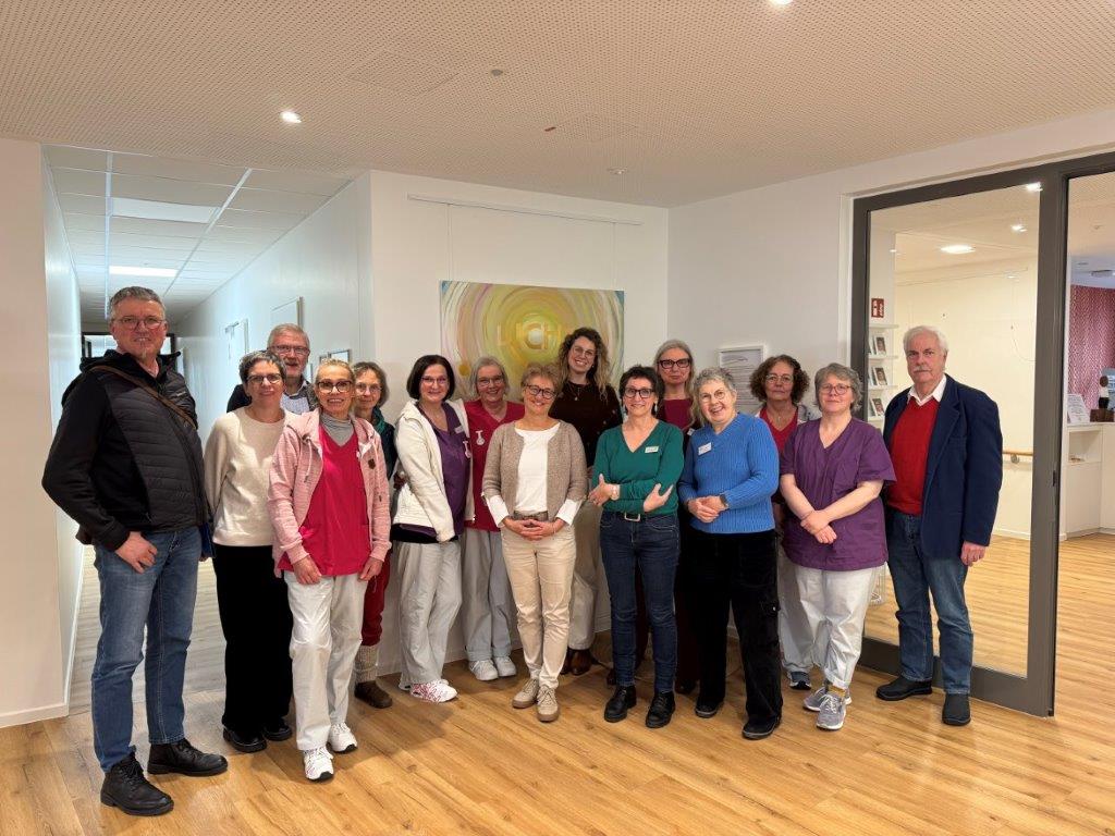 Gruppe von 14 Personen in einem hellen Raum mit Holzboden, einige tragen medizinische Kleidung, andere Freizeitkleidung